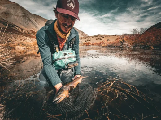 Guide smiling with a trout in the net and the mountains behind
