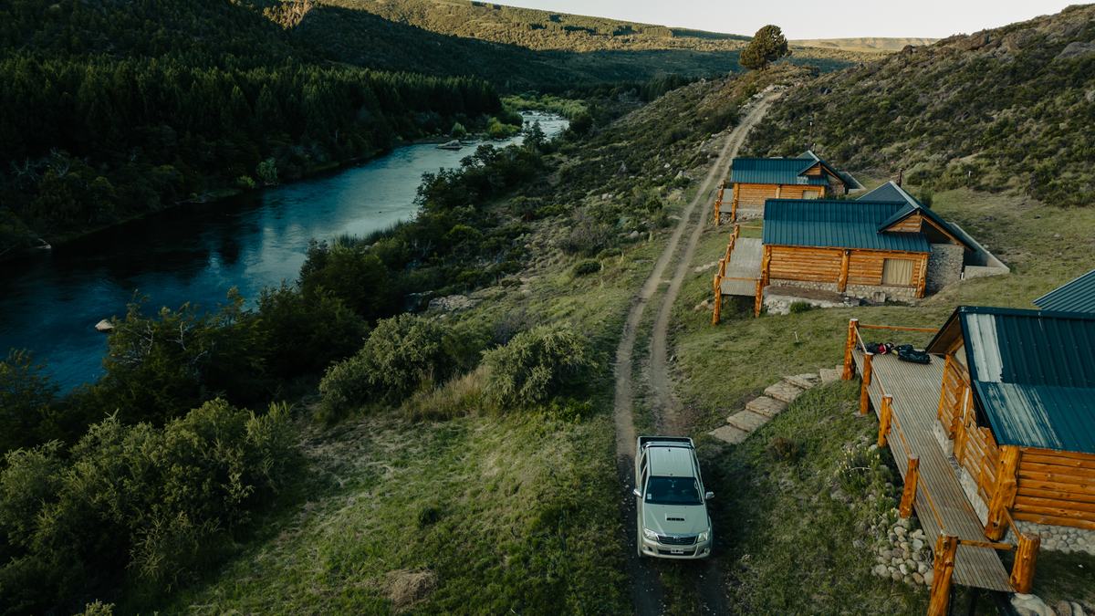 Aerial view of a pickup truck on a dirt track beside log cabins above a river at dusk, northern Patagonia