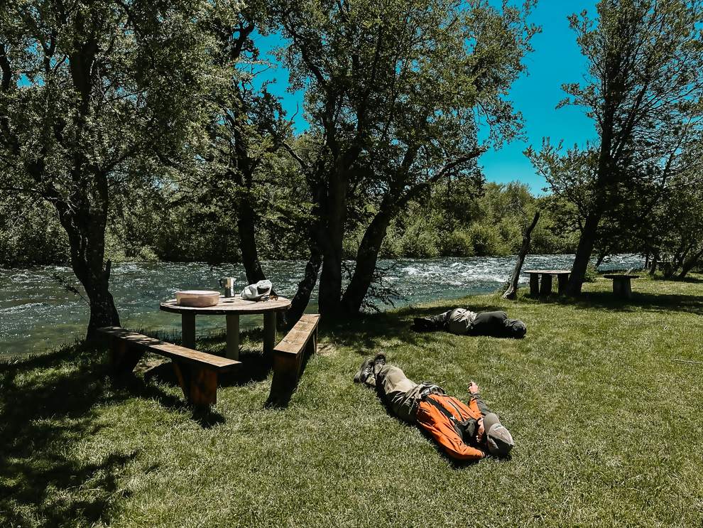 Anglers resting on the grass by the river after lunch under the trees in northern Patagonia